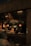 A dimly lit restaurant scene with a focus on a menu board displaying various food items and prices. The interior features hanging light fixtures with warm lighting, giving the space a cozy ambience. Behind the counter, there are glass racks, cooking utensils, and a couple of staff members engaged in work. A patron is seated at a table in the foreground.