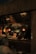 A dimly lit restaurant scene with a focus on a menu board displaying various food items and prices. The interior features hanging light fixtures with warm lighting, giving the space a cozy ambience. Behind the counter, there are glass racks, cooking utensils, and a couple of staff members engaged in work. A patron is seated at a table in the foreground.