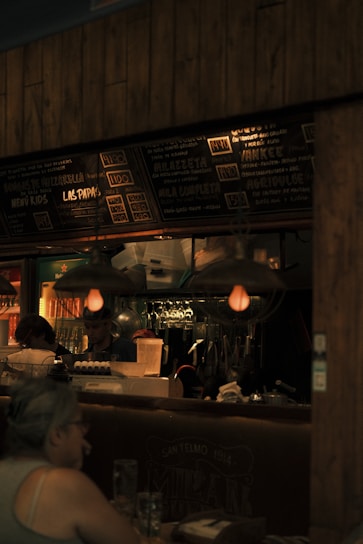 A dimly lit restaurant scene with a focus on a menu board displaying various food items and prices. The interior features hanging light fixtures with warm lighting, giving the space a cozy ambience. Behind the counter, there are glass racks, cooking utensils, and a couple of staff members engaged in work. A patron is seated at a table in the foreground.