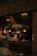 A dimly lit restaurant scene with a focus on a menu board displaying various food items and prices. The interior features hanging light fixtures with warm lighting, giving the space a cozy ambience. Behind the counter, there are glass racks, cooking utensils, and a couple of staff members engaged in work. A patron is seated at a table in the foreground.