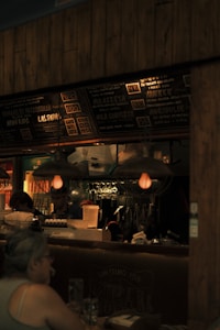 A dimly lit restaurant scene with a focus on a menu board displaying various food items and prices. The interior features hanging light fixtures with warm lighting, giving the space a cozy ambience. Behind the counter, there are glass racks, cooking utensils, and a couple of staff members engaged in work. A patron is seated at a table in the foreground.