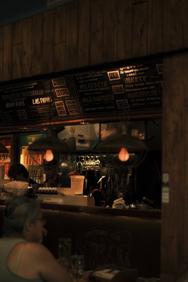 A dimly lit restaurant scene with a focus on a menu board displaying various food items and prices. The interior features hanging light fixtures with warm lighting, giving the space a cozy ambience. Behind the counter, there are glass racks, cooking utensils, and a couple of staff members engaged in work. A patron is seated at a table in the foreground.