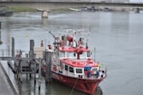 A red and white fire rescue boat is docked at a pier on a calm river. The boat is well-equipped with various equipment and antennas on its deck. There is a person in a yellow jacket standing on the boat's deck. A bridge spans the river in the background, and the surrounding area is urban with visible structures and greenery.