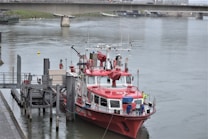 A red and white fire rescue boat is docked at a pier on a calm river. The boat is well-equipped with various equipment and antennas on its deck. There is a person in a yellow jacket standing on the boat's deck. A bridge spans the river in the background, and the surrounding area is urban with visible structures and greenery.