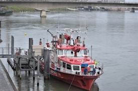 A red and white fire rescue boat is docked at a pier on a calm river. The boat is well-equipped with various equipment and antennas on its deck. There is a person in a yellow jacket standing on the boat's deck. A bridge spans the river in the background, and the surrounding area is urban with visible structures and greenery.