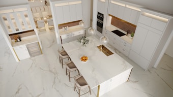 Spacious modern kitchen with white cabinetry and a large marble island at the center. Three pendant lights hang above the island, which houses a sink and is decorated with a bowl of oranges and a flower arrangement. Five bar stools are placed along one side of the island. The kitchen features built-in appliances, including an oven and a refrigerator, with ample lighting and a seamless design.