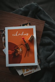 A magazine with an orange cover featuring a lipstick and a wilted flower rests on a wooden surface. The magazine is surrounded by other publications and a shadowy fabric draped behind it, creating a cozy and artistic setting.