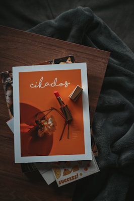 A magazine with an orange cover featuring a lipstick and a wilted flower rests on a wooden surface. The magazine is surrounded by other publications and a shadowy fabric draped behind it, creating a cozy and artistic setting.