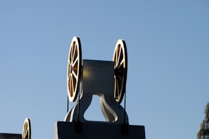 A mechanical structure featuring two large, spoked wheels cast in shadow against a clear blue sky. The wheels appear to be part of an industrial machine or a piece of industrial architecture. The sunlight creates a golden hue on the edges of the wheels, adding a striking contrast to the image.