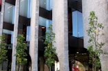 Modern architectural building facade with tall glass windows, stone walls, and several green trees lining the front. The structure features large vertical columns and reflective surfaces, creating a sleek and contemporary appearance.