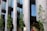 Modern architectural building facade with tall glass windows, stone walls, and several green trees lining the front. The structure features large vertical columns and reflective surfaces, creating a sleek and contemporary appearance.