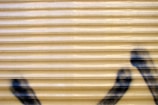 A corrugated metal surface with a beige color is partially spray-painted with black graffiti markings at the bottom right corner.
