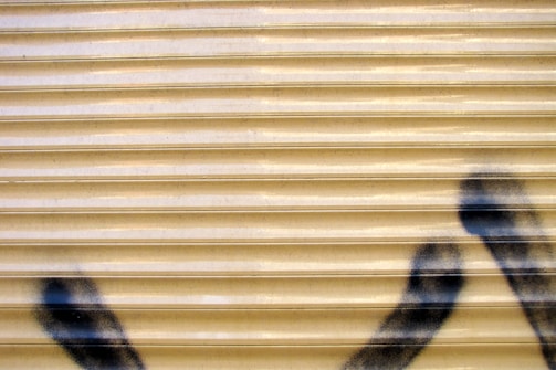 A corrugated metal surface with a beige color is partially spray-painted with black graffiti markings at the bottom right corner.