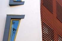 A modern architectural facade featuring asymmetrical windows with dark frames and blue-tinted glass. The wall is white, contrasting with a set of reddish-brown horizontal wooden slats that provide texture and depth.