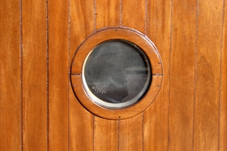 A wooden wall with vertical panels features a round, glass porthole window at the center. The wood has a smooth, polished finish with a rich brown color.