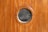 A wooden wall with vertical panels features a round, glass porthole window at the center. The wood has a smooth, polished finish with a rich brown color.