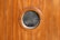 A wooden wall with vertical panels features a round, glass porthole window at the center. The wood has a smooth, polished finish with a rich brown color.