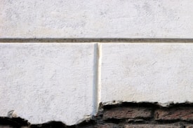 A close-up view of a textured white concrete wall with visible weathering and discoloration. The bottom edge reveals an exposed layer of dark, rough stone or brick, creating a contrast between the smooth, light upper portion and the rugged, darker base.