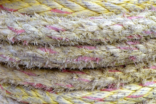 Close-up view of several twisted, frayed, and thick ropes stacked together. The ropes are primarily yellow with interwoven pink strands, giving them a rugged and weathered appearance.