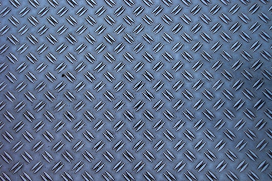 A metal surface with a diamond plate pattern, showing a series of raised, elongated shapes uniformly distributed across the surface.