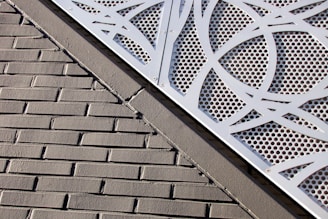 A textured brick wall is intersected by a metal panel featuring a geometric cut-out design with circular perforations. The panel is secured with metal screws at the corners.