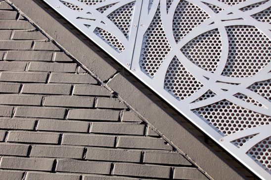 A textured brick wall is intersected by a metal panel featuring a geometric cut-out design with circular perforations. The panel is secured with metal screws at the corners.