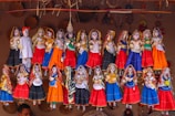 A collection of colorful, traditional Rajasthani puppets, known as kathputlis, are displayed hanging in a row. Each puppet is intricately dressed in vibrant attire, featuring designs of red, blue, orange, and green. The figures are adorned with detailed accessories such as jewelry and headscarves, showcasing cultural craftsmanship. The puppets have expressive facial features painted on them, which adds a lively touch to the display. Above them, a mix of woven straw decorations adds to the rustic cultural theme.