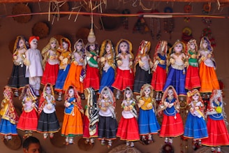 A collection of colorful, traditional Rajasthani puppets, known as kathputlis, are displayed hanging in a row. Each puppet is intricately dressed in vibrant attire, featuring designs of red, blue, orange, and green. The figures are adorned with detailed accessories such as jewelry and headscarves, showcasing cultural craftsmanship. The puppets have expressive facial features painted on them, which adds a lively touch to the display. Above them, a mix of woven straw decorations adds to the rustic cultural theme.
