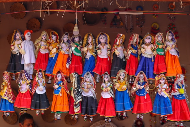 A collection of colorful, traditional Rajasthani puppets, known as kathputlis, are displayed hanging in a row. Each puppet is intricately dressed in vibrant attire, featuring designs of red, blue, orange, and green. The figures are adorned with detailed accessories such as jewelry and headscarves, showcasing cultural craftsmanship. The puppets have expressive facial features painted on them, which adds a lively touch to the display. Above them, a mix of woven straw decorations adds to the rustic cultural theme.