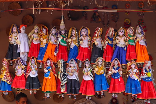 A collection of colorful, traditional Rajasthani puppets, known as kathputlis, are displayed hanging in a row. Each puppet is intricately dressed in vibrant attire, featuring designs of red, blue, orange, and green. The figures are adorned with detailed accessories such as jewelry and headscarves, showcasing cultural craftsmanship. The puppets have expressive facial features painted on them, which adds a lively touch to the display. Above them, a mix of woven straw decorations adds to the rustic cultural theme.