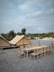 A campsite featuring two beige tents set up on wooden platforms amidst a gravel ground. In front of the tents, there is a long wooden table surrounded by several matching wooden chairs. The background includes a few trees and some cloudy skies, indicating a tranquil outdoor setting.