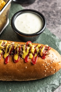 A sesame seed hot dog bun is filled with seasoned meat and topped with lines of red ketchup and yellow mustard. Beside it, a small black ceramic cup contains a creamy white sauce. The surface is textured, resembling slate, providing a rustic background.