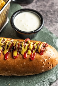 A sesame seed hot dog bun is filled with seasoned meat and topped with lines of red ketchup and yellow mustard. Beside it, a small black ceramic cup contains a creamy white sauce. The surface is textured, resembling slate, providing a rustic background.