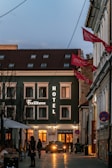 A cozy urban street scene during dusk featuring a hotel on the left side, illuminated by warm, glowing lights. The facade of the hotel is dark with several windows and signs reading 'Hotel' and 'Fulton.' A car is parked in front, and a few pedestrians, are walking along the street, adding to the lively atmosphere. Red flags hang from the building on the right, flapping in the gentle breeze.