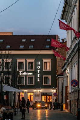 A cozy urban street scene during dusk featuring a hotel on the left side, illuminated by warm, glowing lights. The facade of the hotel is dark with several windows and signs reading 'Hotel' and 'Fulton.' A car is parked in front, and a few pedestrians, are walking along the street, adding to the lively atmosphere. Red flags hang from the building on the right, flapping in the gentle breeze.