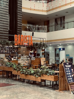 A modern indoor coffee shop located in a shopping mall with a prominent 'BARISTA' sign. The shop features wooden furniture, a counter with a glass display of pastries, potted plants, and a safety signboard. The area is well-lit with overhead lighting and natural light from the mall's upper level. The background shows various other storefronts.