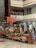 A modern indoor coffee shop located in a shopping mall with a prominent 'BARISTA' sign. The shop features wooden furniture, a counter with a glass display of pastries, potted plants, and a safety signboard. The area is well-lit with overhead lighting and natural light from the mall's upper level. The background shows various other storefronts.