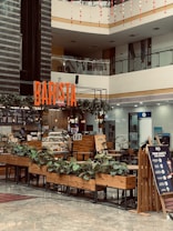 A modern indoor coffee shop located in a shopping mall with a prominent 'BARISTA' sign. The shop features wooden furniture, a counter with a glass display of pastries, potted plants, and a safety signboard. The area is well-lit with overhead lighting and natural light from the mall's upper level. The background shows various other storefronts.