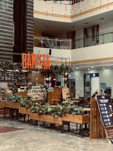 A modern indoor coffee shop located in a shopping mall with a prominent 'BARISTA' sign. The shop features wooden furniture, a counter with a glass display of pastries, potted plants, and a safety signboard. The area is well-lit with overhead lighting and natural light from the mall's upper level. The background shows various other storefronts.