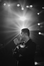 A black and white portrait of a man standing with his arms crossed, gazing thoughtfully to the side. Bright stage lights shine in the background, creating a dramatic effect with rays spreading outwards.