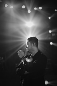 A black and white portrait of a man standing with his arms crossed, gazing thoughtfully to the side. Bright stage lights shine in the background, creating a dramatic effect with rays spreading outwards.