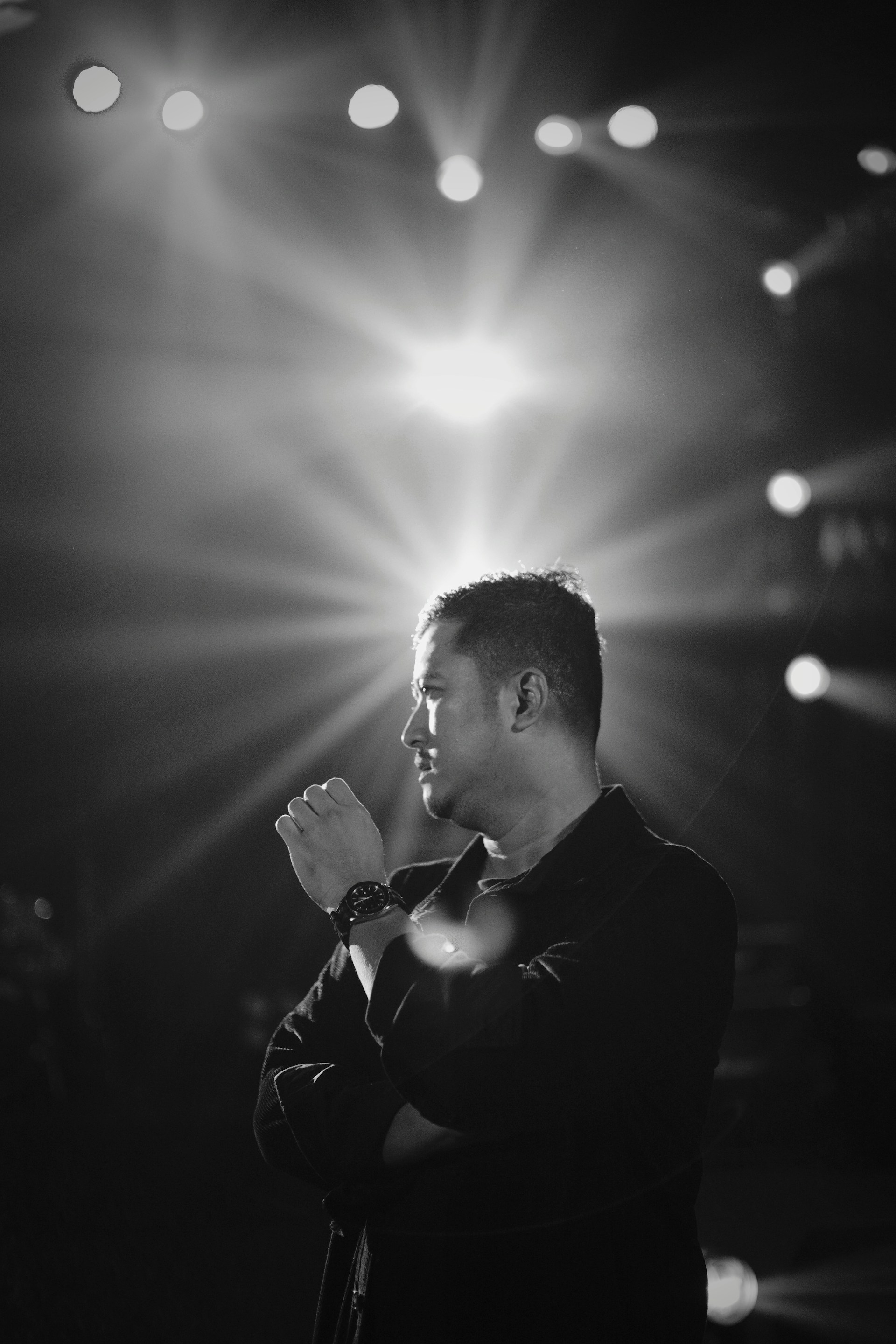 A black and white portrait of a man standing with his arms crossed, gazing thoughtfully to the side. Bright stage lights shine in the background, creating a dramatic effect with rays spreading outwards.