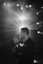 A black and white portrait of a man standing with his arms crossed, gazing thoughtfully to the side. Bright stage lights shine in the background, creating a dramatic effect with rays spreading outwards.