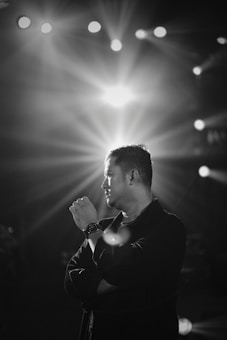 A black and white portrait of a man standing with his arms crossed, gazing thoughtfully to the side. Bright stage lights shine in the background, creating a dramatic effect with rays spreading outwards.