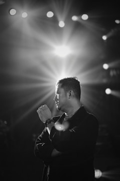 A black and white portrait of a man standing with his arms crossed, gazing thoughtfully to the side. Bright stage lights shine in the background, creating a dramatic effect with rays spreading outwards.