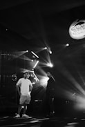 Two people stand in a dimly lit setting, with stage lights creating dramatic rays and shadows. A logo or sign hangs in the upper right corner, reading 'Collab...' which suggests a musical or entertainment event.