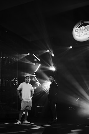Two people stand in a dimly lit setting, with stage lights creating dramatic rays and shadows. A logo or sign hangs in the upper right corner, reading 'Collab...' which suggests a musical or entertainment event.