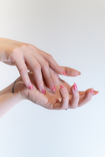 Two hands are positioned elegantly against a neutral background, with nails painted in a gradient of pink shades featuring intricate designs. The hands convey a sense of graceful movement and creativity.