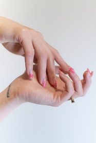 Two hands are positioned in a graceful, intertwined pose, displaying an artistic nail design with pink, flame-like patterns. A small tattoo is visible on the wrist, and a ring adorns one finger.