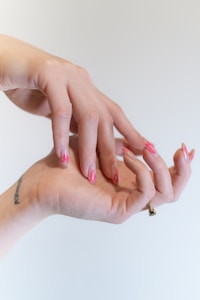 Two hands are positioned in a graceful, intertwined pose, displaying an artistic nail design with pink, flame-like patterns. A small tattoo is visible on the wrist, and a ring adorns one finger.